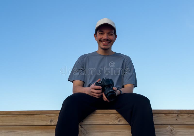 Fotógrafo sonriente sentado en una estructura de madera con un cielo azul en el fondo fotografía de archivo