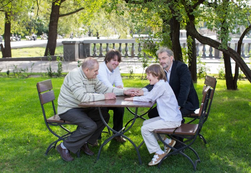 Four generations of men sitting at a wooden table in a park, laughing and talking