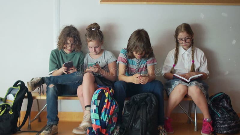 Four schoolchildren sit on a bench at school and look at the screens of their smartphones. One girl with glasses is