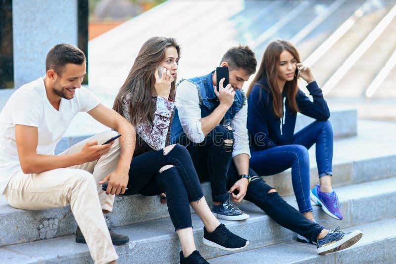 Four young people sitting on stairs and using mobile phones. Two women and one men talking on smartphone and one guy browsing. Modern life and technology concept