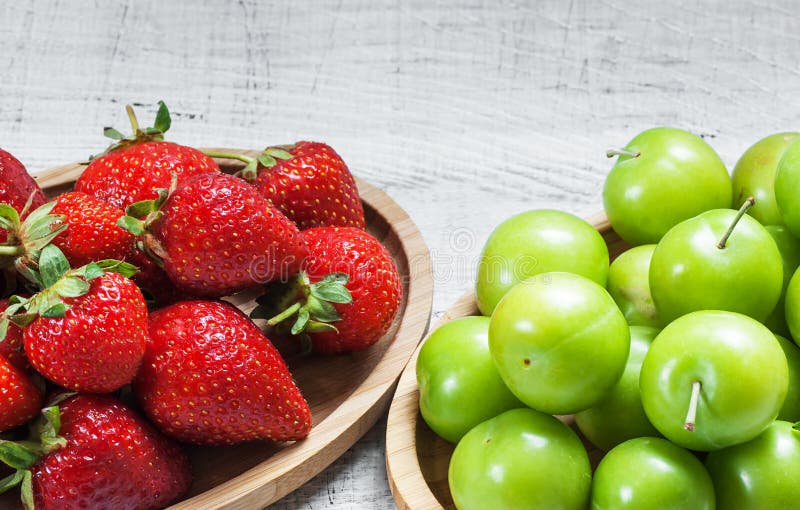 Fraises fraîches mûres et prunes vertes dans la cuvette en bambou sur le fond en bois blanc images stock Fraises fraîches mûres et prunes vertes dans la cuvette en bambou sur le fond en bois blanc images stock