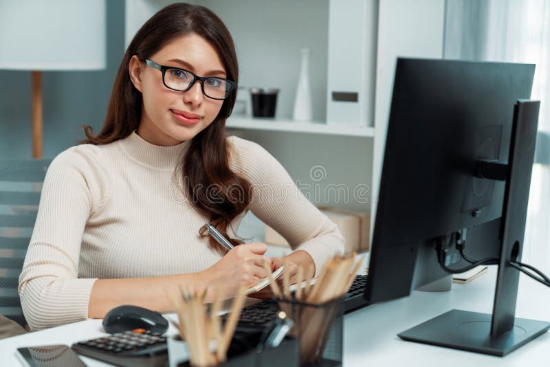 Portrait casual outfit smiling beautiful woman looking camera to pose for business profile while working on desk surrounded pc and stationary theme of positive new generation modern office. Postulate. Portrait casual outfit smiling beautiful woman looking camera to pose for business profile while working on desk surrounded pc and stationary theme of positive new generation modern office. Postulate.