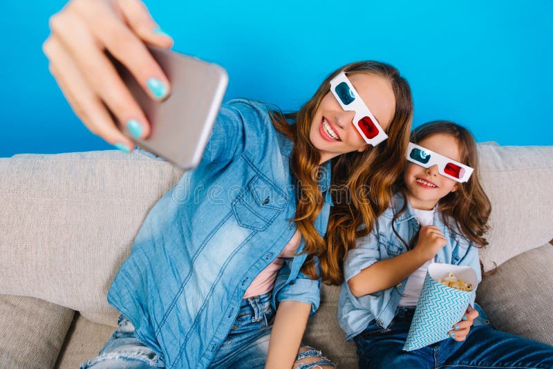Framställning av den lyckliga selfieståenden av den nätta unga modern med den glade dottern som ler till kameran på soffan på blå arkivfoton Framställning av den lyckliga selfieståenden av den nätta unga modern med den glade dottern som ler till kameran på soffan på blå arkivfoton
