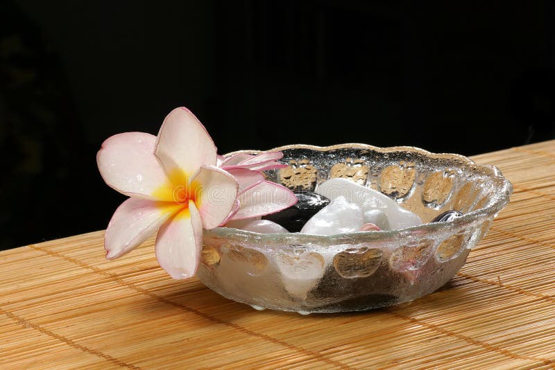 Detail of frangipane flower and pebbles in a glass bowl on the rattan background. Detail of frangipane flower and pebbles in a glass bowl on the rattan background