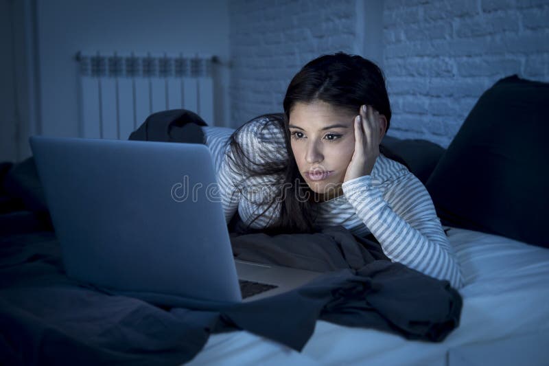 Young and beautiful hispanic woman in pajamas on bed at home bedroom working bored and tired with laptop computer late at night in dark room light looking sleepy. Young and beautiful hispanic woman in pajamas on bed at home bedroom working bored and tired with laptop computer late at night in dark room light looking sleepy