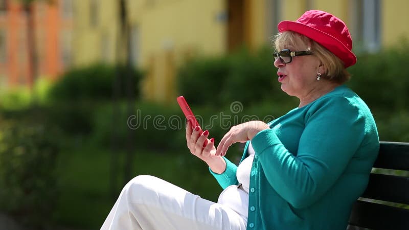 Frau in der roten Kappe sitzt auf der Bank und steht über Smartphone in Verbindung stock footage Frau in der roten Kappe sitzt auf der Bank und steht über Smartphone in Verbindung stock footage