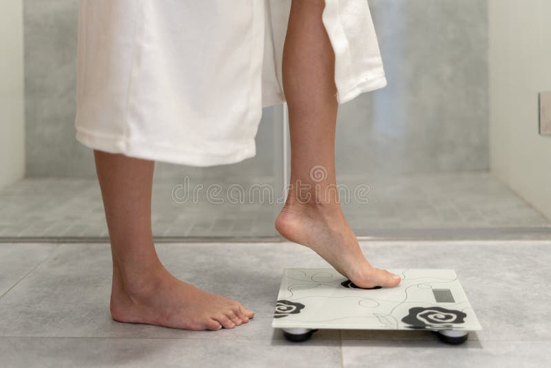 Cropped view of woman in white and comfort bathrobe making step on weigher in bathroom with copy space. Cropped view of woman in white and comfort bathrobe making step on weigher in bathroom with copy space