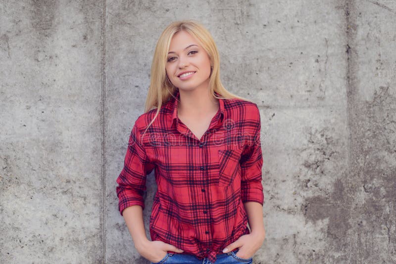 Woman smiling at camera. Woman with blonde hair, dressed in red checkered shirt and jeans standing against grey wall. Portrait, close up photo. Woman smiling at camera. Woman with blonde hair, dressed in red checkered shirt and jeans standing against grey wall. Portrait, close up photo