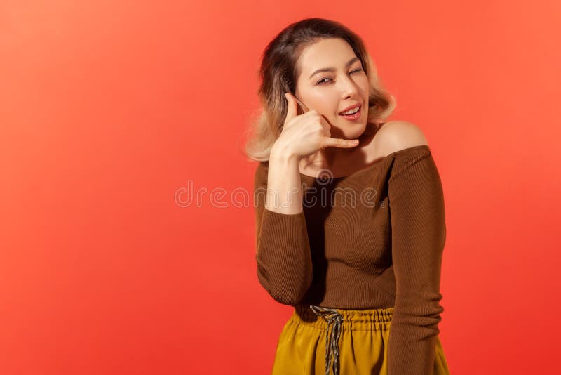 Portrait of young pretty woman in brown casual blouse smiling, showing a gesture of a phone and asking for call her later or telling she will call later. Indoor studio shot isolated on red background. Portrait of young pretty woman in brown casual blouse smiling, showing a gesture of a phone and asking for call her later or telling she will call later. Indoor studio shot isolated on red background