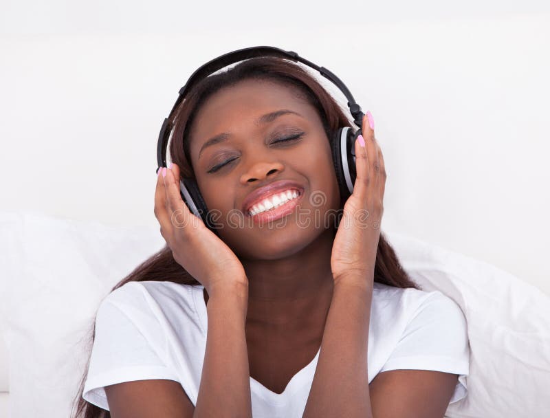 Happy African American woman enjoying music through headphones in bed at home. Happy African American woman enjoying music through headphones in bed at home