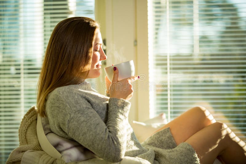 Frau, die zu Hause in einem Stuhl am Fenster mit Schale heißem Kaffee sitzt