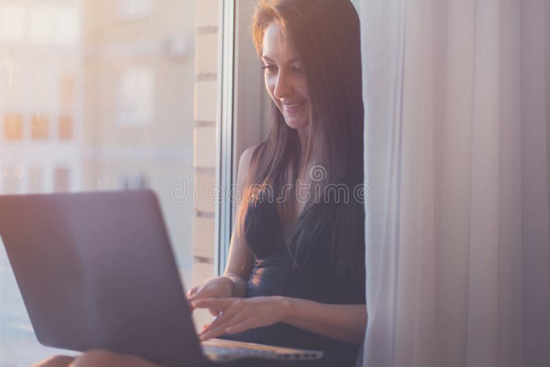 Frau, die zu Hause die Laptop-Computer sitzt nahe Fenster lächelt und verwendet stockbild Frau, die zu Hause die Laptop-Computer sitzt nahe Fenster lächelt und verwendet stockbild