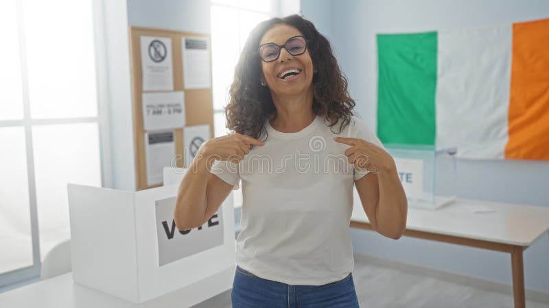 Frau lächelt drinnen in einem Wahlstudienzimmer mit irischer Flagge, die auf sich selbst zeigt, mit Stimmzeichen im Hintergrund lizenzfreies stockfoto