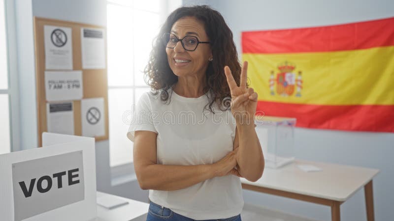 Frau lächelt drinnen und zeigt Friedenszeichen in der spanischen Wahlstation mit spain flagge im Hintergrund lizenzfreies stockfoto