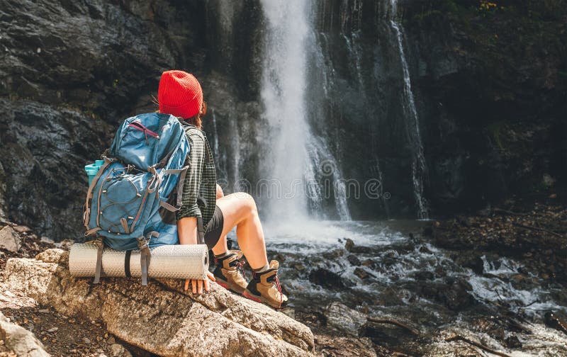 Frau mit einem Rucksack in einem roten Hut in aktiven Trekkingbekleidung, sitzend am Wasserfall des Gebirges und genießt den lizenzfreie stockbilder