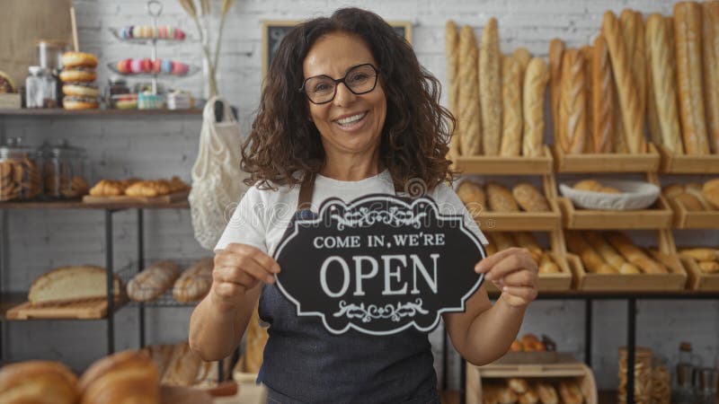 Frau mit offenem Schild in Bäckerei umgeben von Brot und Gebäck lächelnd reife hispanic weibliche Gäste willkommen zu stockfotografie
