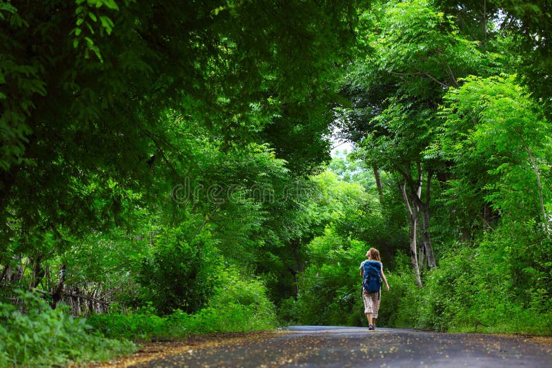 Woman with backpack walking on an asphalt road in a park with green trees. Woman with backpack walking on an asphalt road in a park with green trees