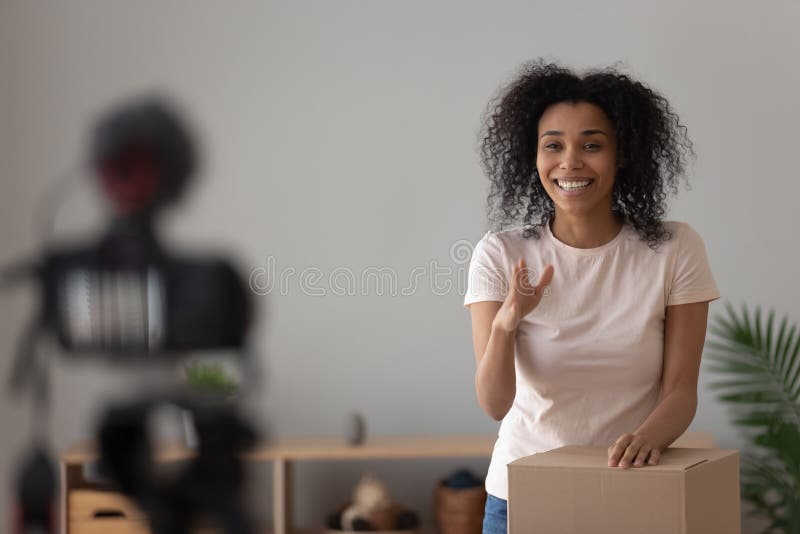 African woman vlogger record new vlog about product or goods, ready unpack package capture moment of opening long awaited cardboard box and filming it on DSLR digital camera out of focus on foreground. African woman vlogger record new vlog about product or goods, ready unpack package capture moment of opening long awaited cardboard box and filming it on DSLR digital camera out of focus on foreground