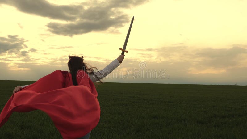 Free girl in a red cloak runs with swords in his hand across field playing medieval knight. young girl playing super