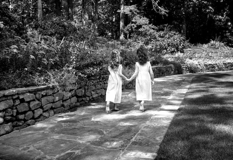 Two young girls skip along a garden path as they enjoy the freedom of just being kids. Both have on dresses and have long curly hair. Black and white photo. Two young girls skip along a garden path as they enjoy the freedom of just being kids. Both have on dresses and have long curly hair. Black and white photo.