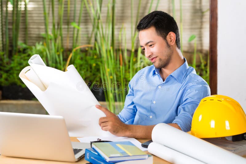 Freelancer - Architect working at home on a design or draft, on his desk are books, a laptop and a helmet or hard hat. Freelancer - Architect working at home on a design or draft, on his desk are books, a laptop and a helmet or hard hat