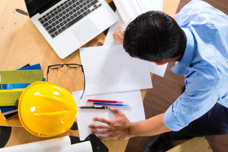 Freelancer - Architect working at home on a design or draft, on his desk are books, a laptop and a helmet or hard hat. Freelancer - Architect working at home on a design or draft, on his desk are books, a laptop and a helmet or hard hat