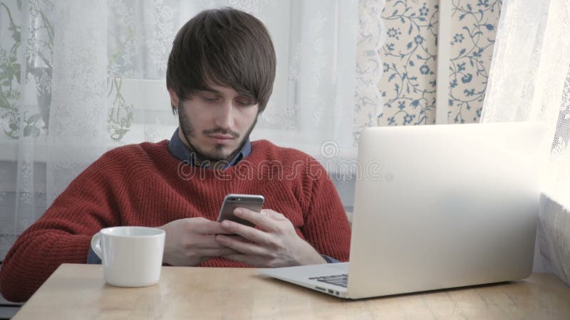 Freelancer feliz do homem novo que trabalha com portátil e o telefone modernos no café que toma a xícara de café