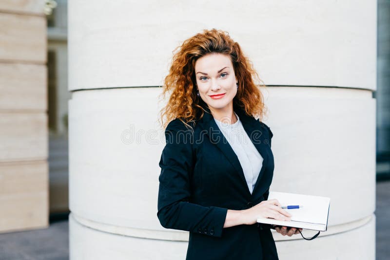 Freelancer fêmea com penteado espesso, a blusa branca vestindo, o revestimento preto e a saia, guardando seu livro do diário, esc fotografia de stock royalty free Freelancer fêmea com penteado espesso, a blusa branca vestindo, o revestimento preto e a saia, guardando seu livro do diário, esc fotografia de stock royalty free