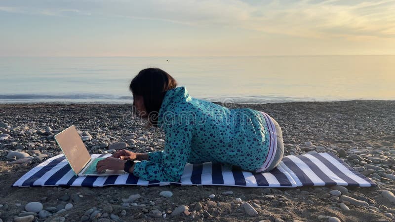 Freelancer fêmea digita mensagem em um laptop portátil e fica na praia. nômada digital. mulher durante o outono