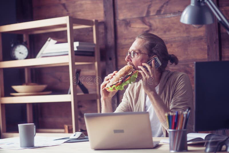 Freelancer que come o sanduíche ao falar no telefone foto de stock Freelancer que come o sanduíche ao falar no telefone foto de stock