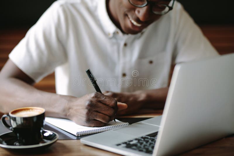 Man writing notes sitting at a coffee shop with a laptop on the