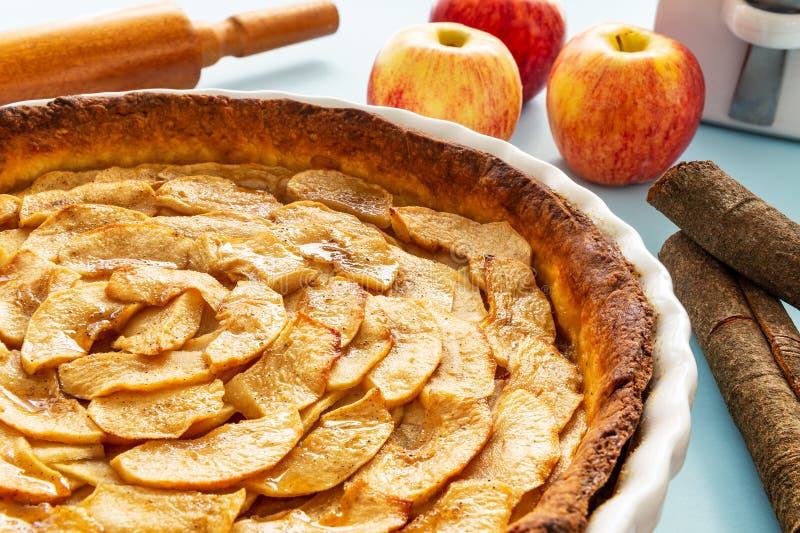 French apple tart aside Gala apples, cinnamon sticks and a rolling pin on a light blue background. Side view