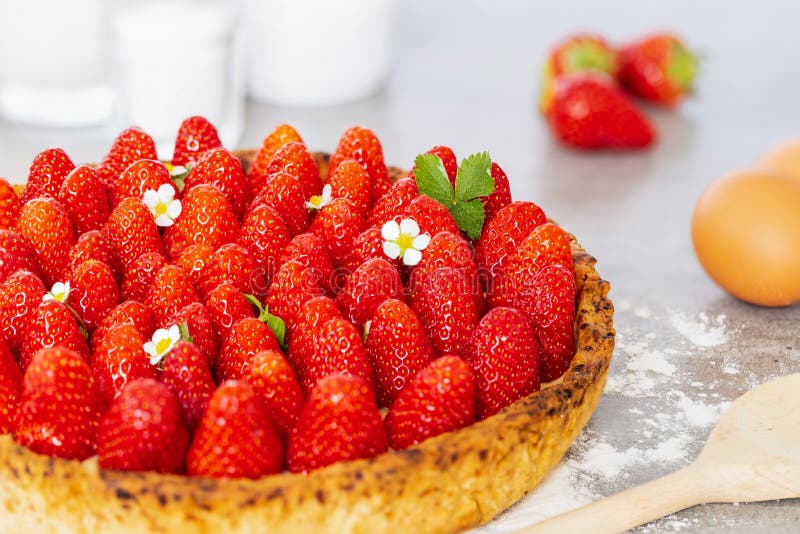 French strawberry tart with wild strawberry leaves and flowers with sugar, spoon and eggs on a grey background with flour stock photography French strawberry tart with wild strawberry leaves and flowers with sugar, spoon and eggs on a grey background with flour stock photography