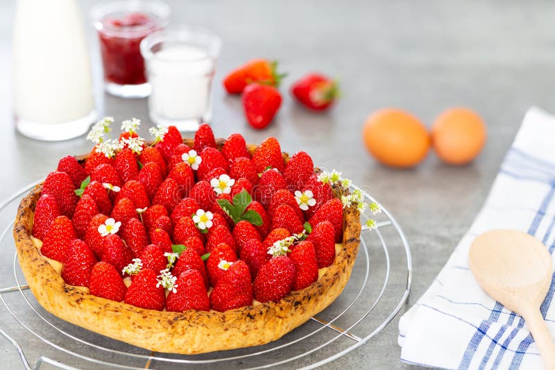 French strawberry tart with wild strawberry leaves and flowers on top of a cooling rack with sugar, jam, spoon and eggs on grey stock image French strawberry tart with wild strawberry leaves and flowers on top of a cooling rack with sugar, jam, spoon and eggs on grey stock image