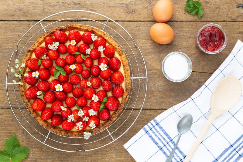 French strawberry tart with wild strawberry leaves and flowers on top on a stainless cooling rack, eggs jam and spoons on wood royalty free stock photos French strawberry tart with wild strawberry leaves and flowers on top on a stainless cooling rack, eggs jam and spoons on wood royalty free stock photos