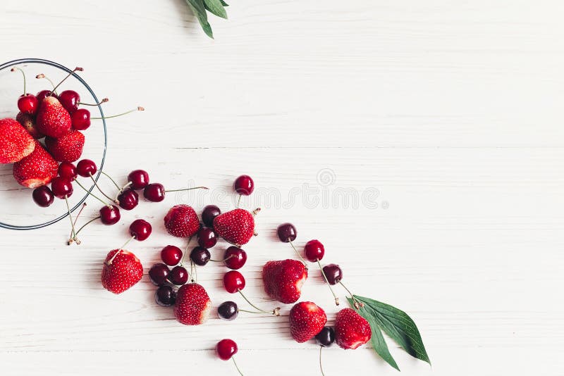 fresh cherries and strawberries scattered from bowl on white rustic wooden background. ripe juicy red berries on table, harvest