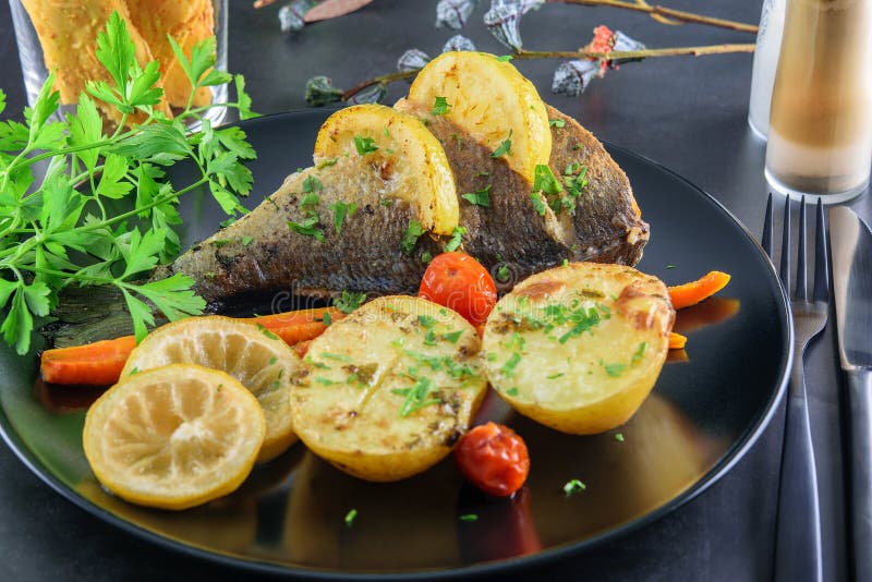 Fresh fried fish dorada with fried slices of potatoes, carrot, tomatoes cherry, lemon and parsley on the black modern dish, served stock photos