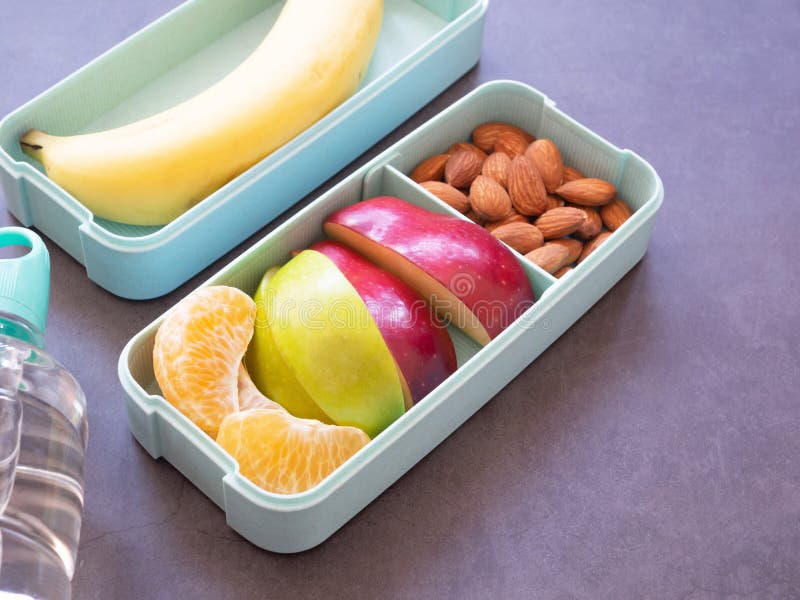 Fresh fruit in light green lunch box with almond, apple, orange, banana, water bottle on grey background. Take away food.