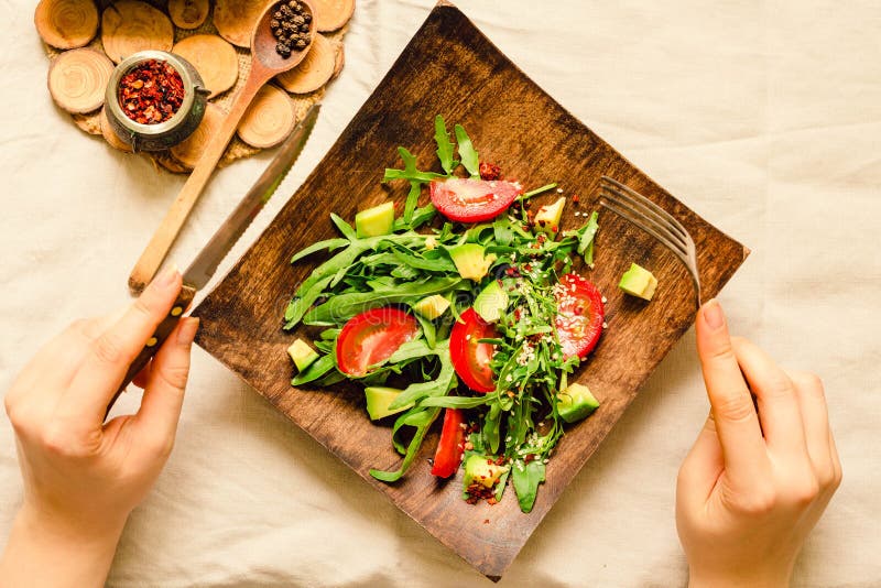 Fresh light green salad with avocado and tomatoes in a wooden plate. Top view. The process of eating. Hands of a young woman stock images Fresh light green salad with avocado and tomatoes in a wooden plate. Top view. The process of eating. Hands of a young woman stock images