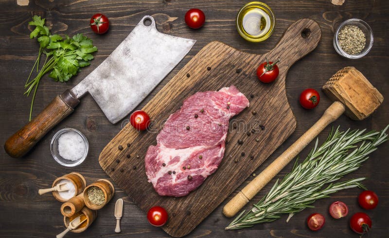 Fresco bistecca di maiale su un tagliere con il rosmarino, un martello per battere la carne e ax per la carne, condimenti ed erbe di legno rustico vista dall'alto.