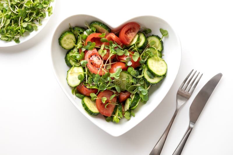 Fresh salad with tomato, cucumber, vegetables, microgreen radishes in plate shape of heart on white. Top view. Concept vegan and