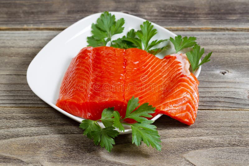 Closeup horizontal photo of fresh red salmon fillet on white plate with parsley on the side and rustic wood underneath. Closeup horizontal photo of fresh red salmon fillet on white plate with parsley on the side and rustic wood underneath