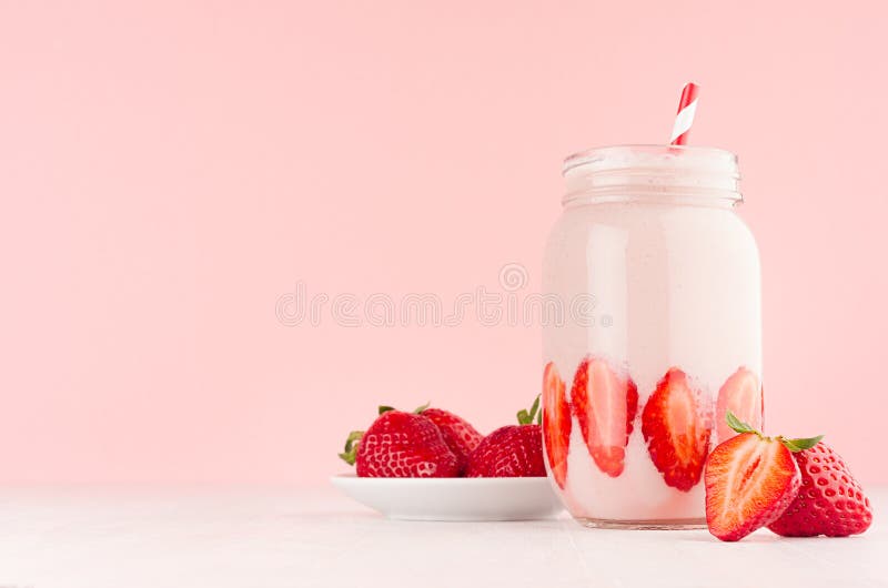 Fresh strawberry milk dessert for breakfast in jar with ripe berry on saucer, straw on pastel pink wall and white wooden table.