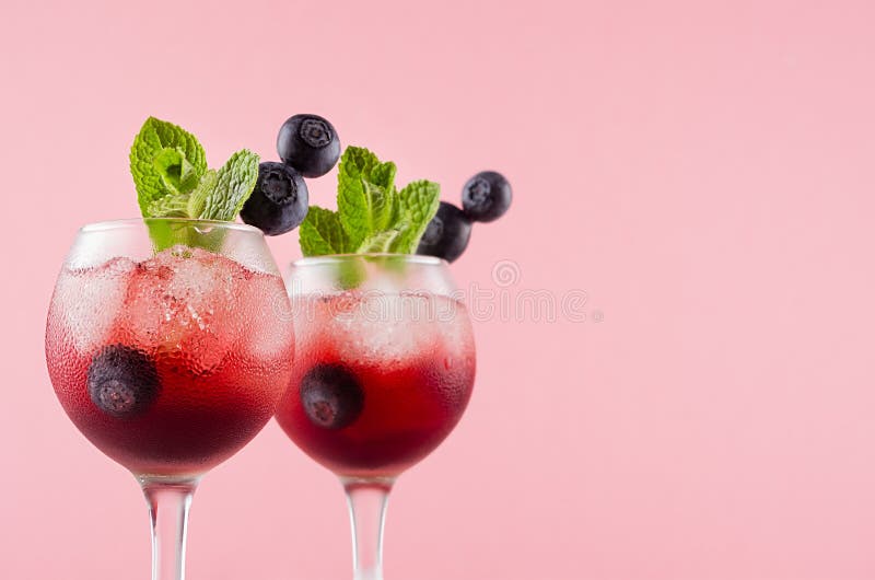 Fresh summer juicy cocktails in two goblets with ice cubes, blueberry, green mint in modern youth pastel soft pink color.