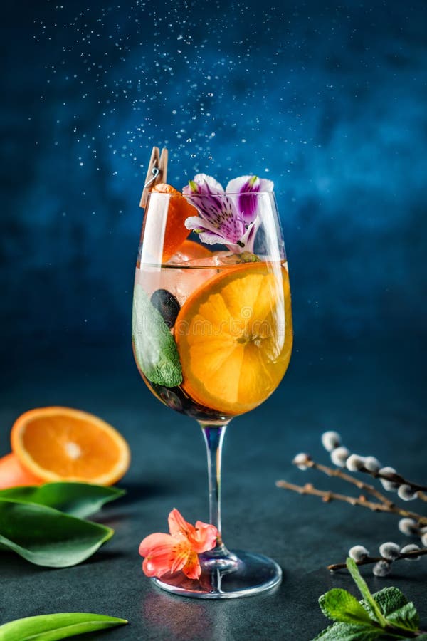 Fresh tropical cocktail with orange, grapes, mint, edible flowers and ice in wine glass on dark blue background