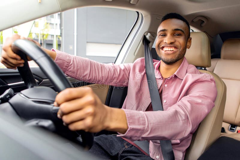 Freudlicher afrikanischer Amerikaner, der Auto fährt und beim Kameralächeln mit Sicherheitsgurt sitzt und Händen hält lizenzfreie stockfotografie