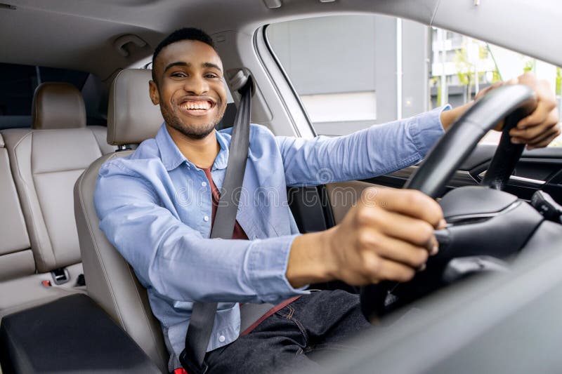 Freudlicher afrikanischer Amerikaner, der Auto fährt und beim Kameralächeln mit Sicherheitsgurt sitzt und Händen hält stockbilder