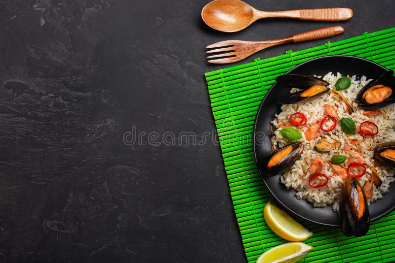 Fried rice with seafood mussels, shrimps, basil in a black plate with wooden spoon and fork on green bamboo mat and stone table