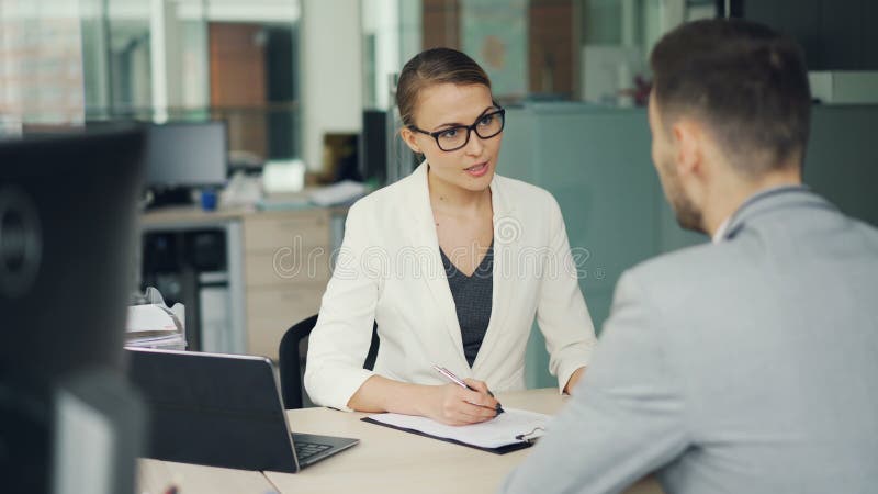 Friendly businesswoman in glasses and suit is interviewing a male candidate for job in office. People are talking asking