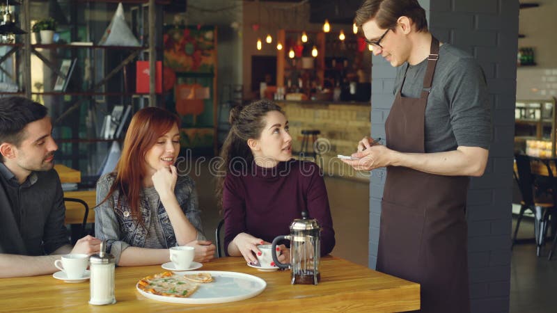 Friendly male waiter is taking order from cheerful group of friends sitting at table in cafe and talking. Eating out